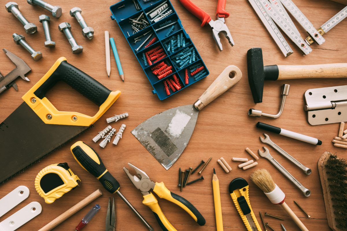 Assorted hand tools and hardware arranged on wooden surface including hammer, pliers, screwdriver, measuring tape, nails, and bolts